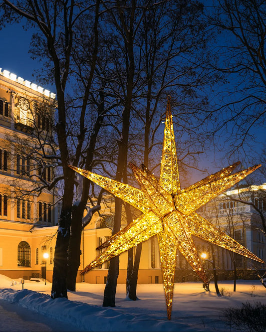 Großer, 12zackiger, gold leuchtender Stern aus Alu mit integrierten LED-Lichtern, aufgestellt im verschneiten Stadtpark vor historischen Gebäuden am Abendhimmel mit blauen Lichtverhältnissen und kahlen Bäumen.