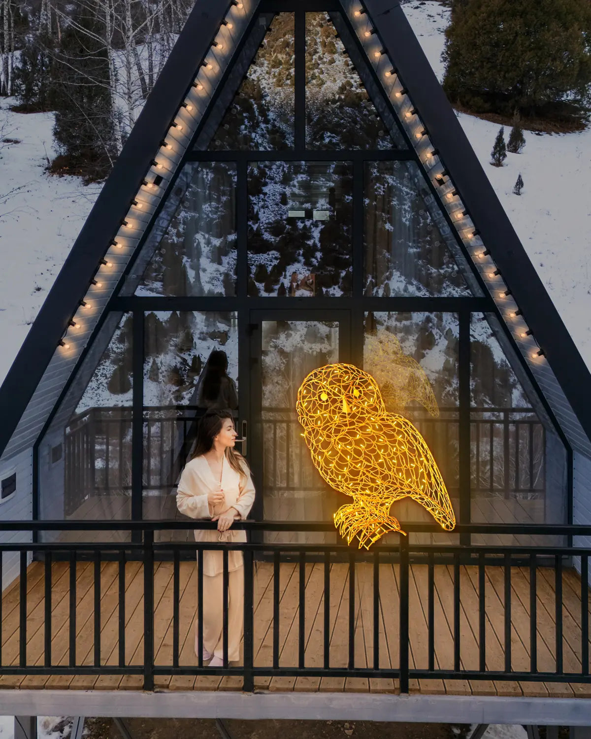 Leuchtende Eulen-Skulptur auf dem Balkon eines modernen Holzhauses im Winter – warme Lichtakzente vor verschneiter Berglandschaft.