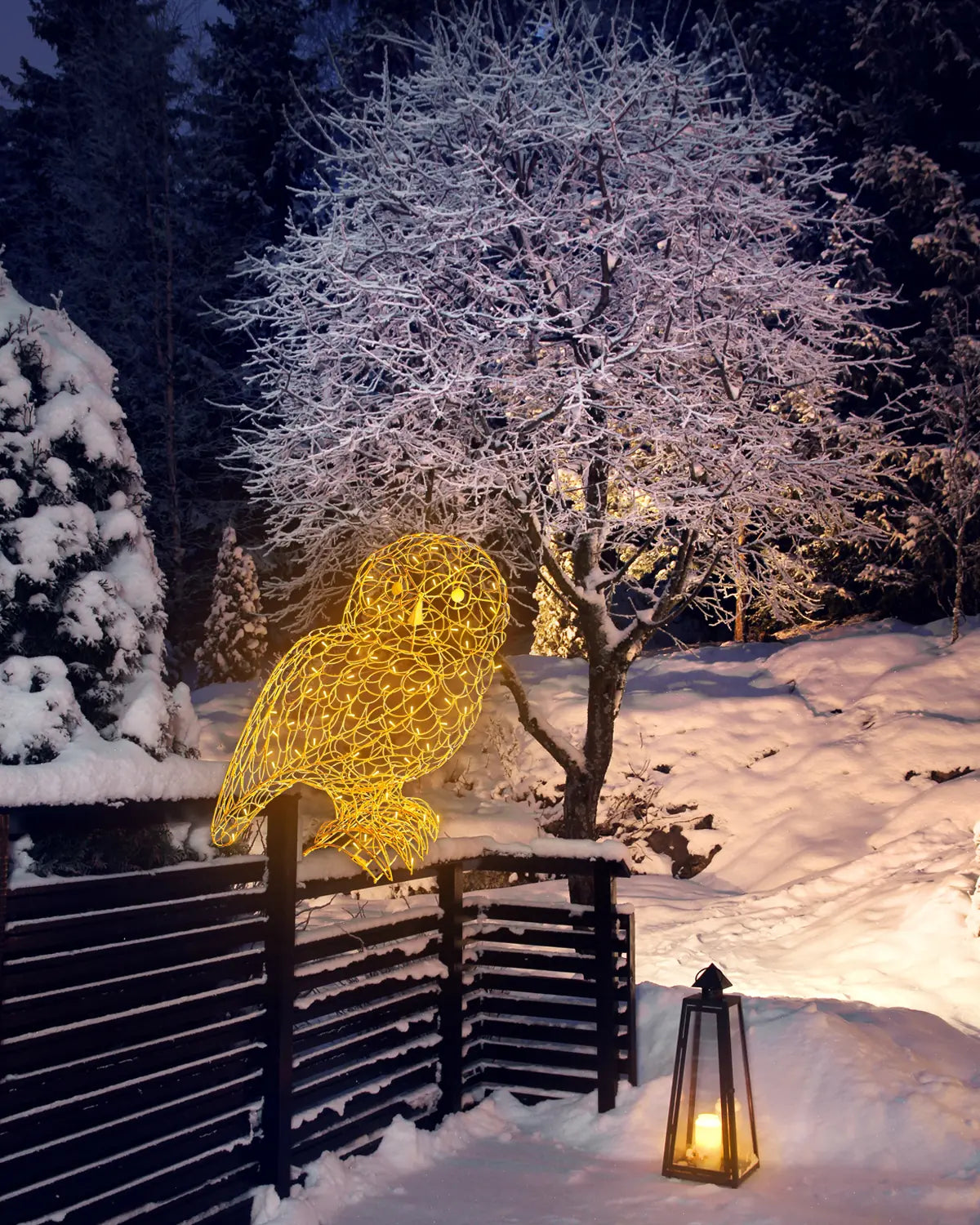Leuchtende Eulen-Skulptur aus goldenem Draht auf einem verschneiten Holzzaun bei Nacht, mit winterlicher Waldkulisse, schneebedeckten Bäumen und einer brennenden Laterne im Vordergrund.