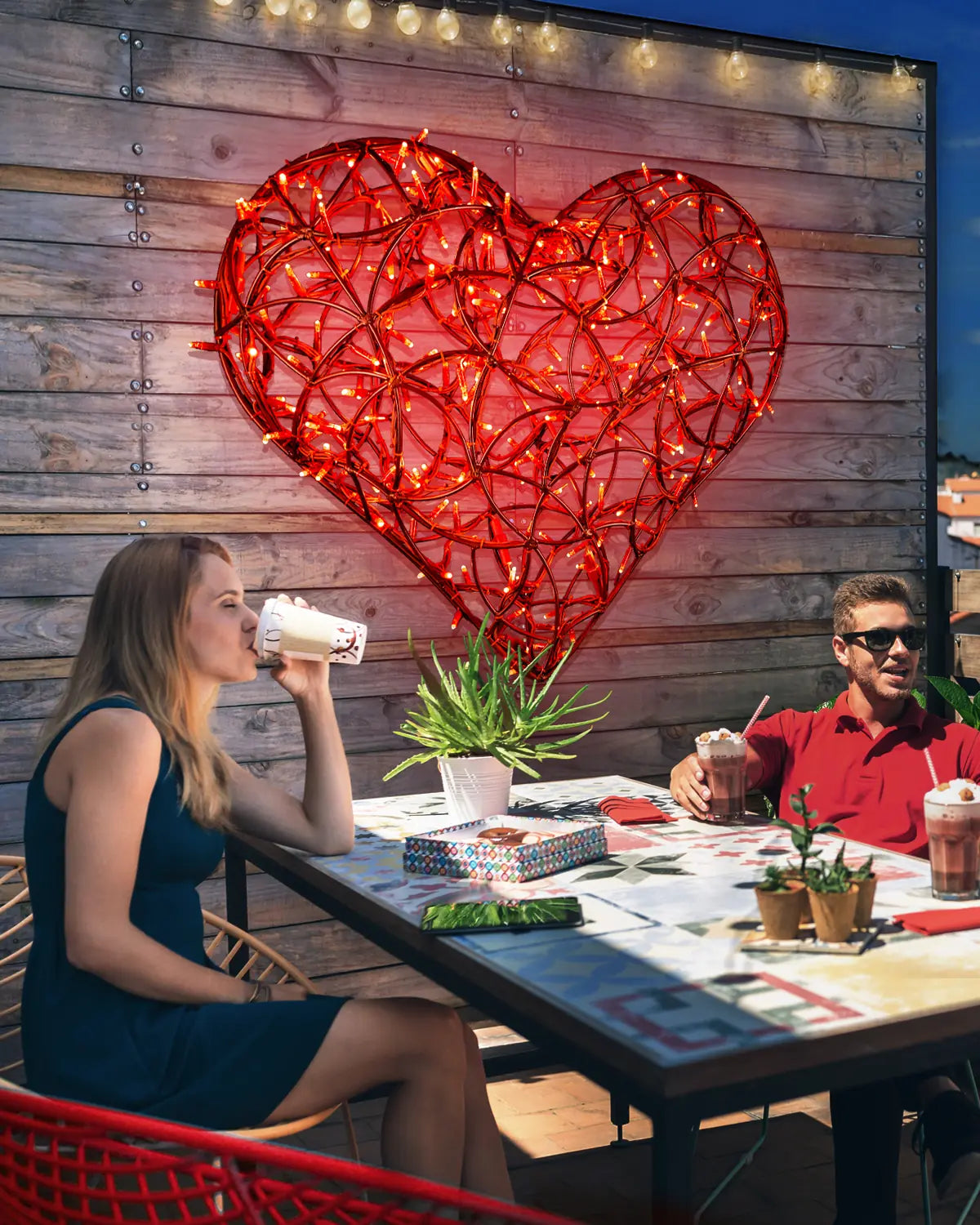 Zwei Personen sitzen an einem bunt gedeckten Tisch auf einer Terrasse, während ein großes, leuchtend rotes Herz aus Lichtschläuchen an einer Holzwand im Hintergrund befestigt ist. Die Szene wirkt sommerlich und einladend.