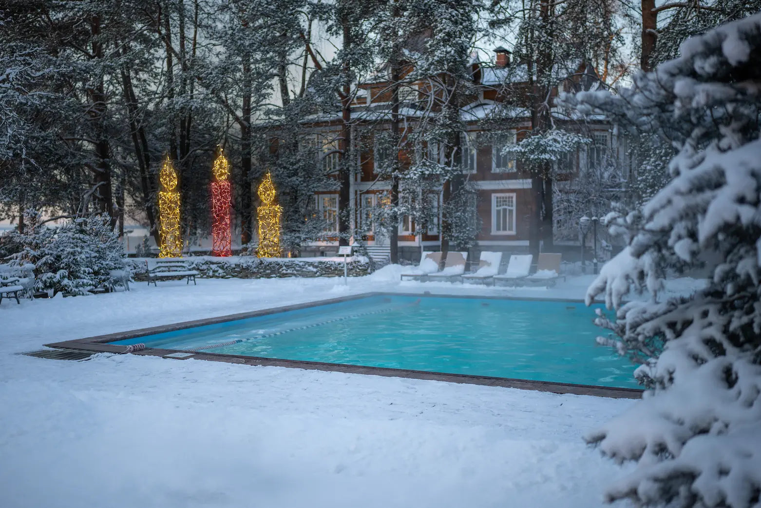 Winterliche Außenaufnahme eines verschneiten Hotelgartens mit beleuchtetem Pool und drei großen leuchtenden Weihnachtskerzen-Dekorationen im Hintergrund – festliche Stimmung trotz Schnee.