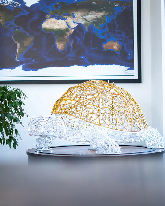 Große Lichtskulptur in Schildkrötenform aus gold-weißer Aluminiumstruktur – inszeniert im Innenraum vor Weltkarte.