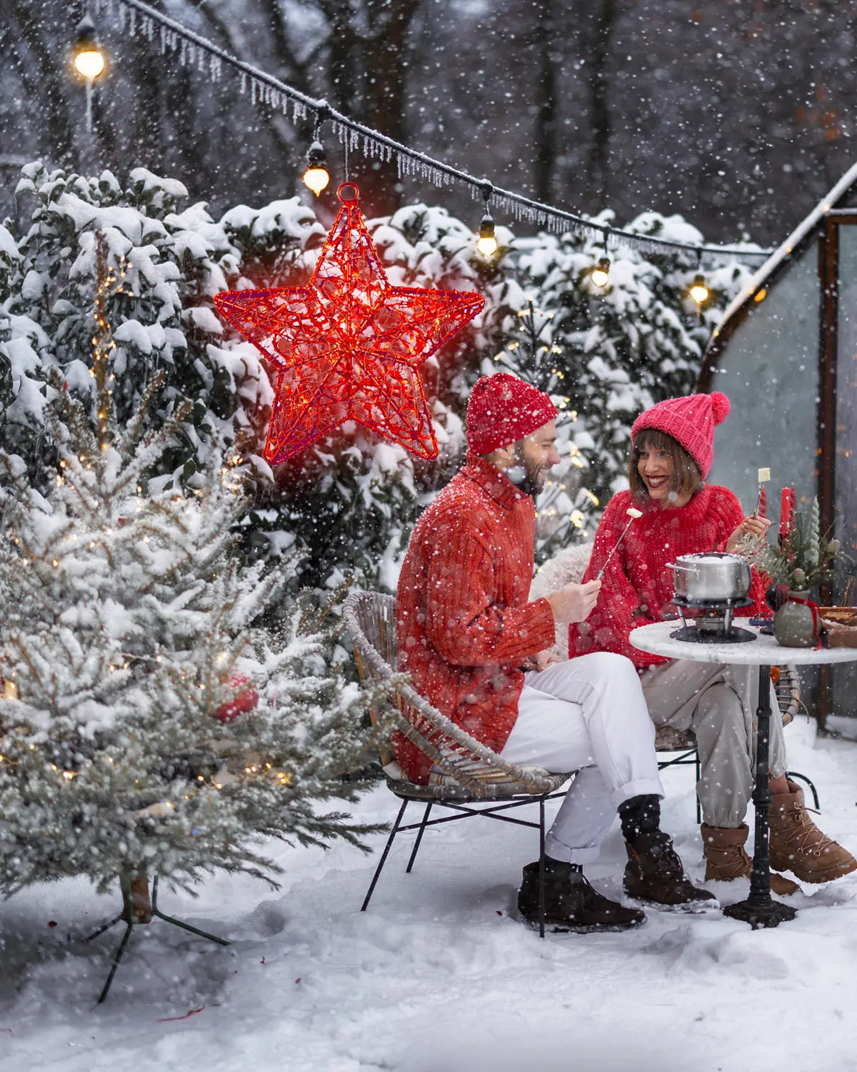 Paar sitzt im verschneiten Garten bei Fondue unter einer Lichterkette mit rotem Stern – gemütliche Winterstimmung mit Weihnachtsbeleuchtung im Außenbereich.