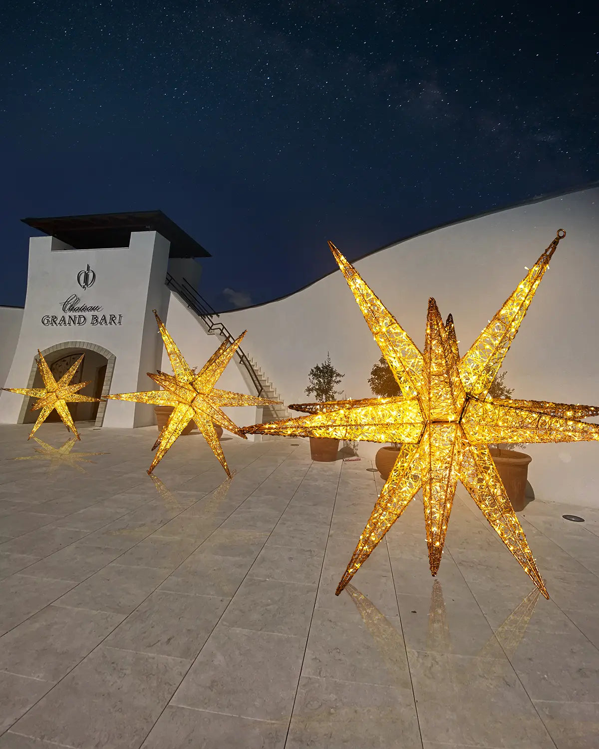 Großformatige Alganic-Sterne auf Marmorterrasse vor moderner Architektur – goldene Lichtinstallation bei Nacht vor dem Château Grand Bari.