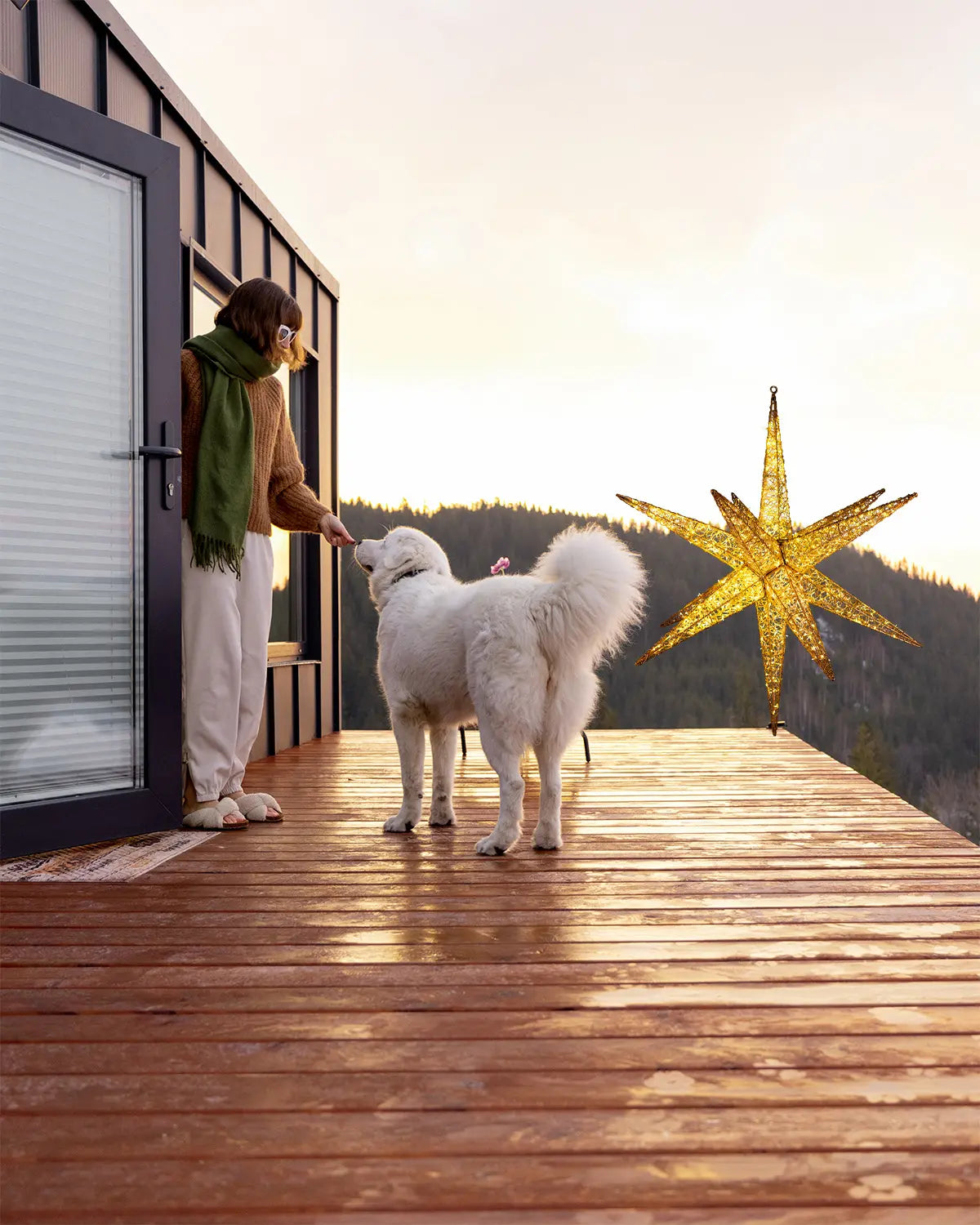 Frau streichelt weißen Hund auf Holzterrasse, daneben eine große hängende goldene Sternfigur, Hintergrund mit bewaldeten Hügeln bei Sonnenuntergang.