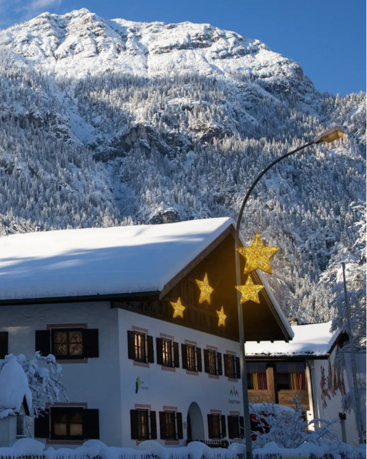 Leuchtsterne von Alganic an einem verschneiten Haus und Laternenmast im winterlichen Alpenpanorama – festliche Außenbeleuchtung mit goldenen Sternen vor Bergkulisse.