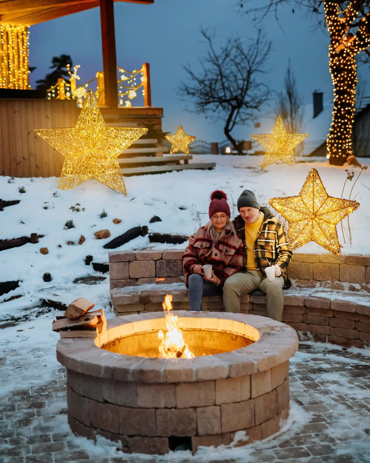 Goldene Lichtsterne als Outdoor-Dekoration rund um eine Feuerstelle im verschneiten Garten – winterliche Abendstimmung mit gemütlicher Atmosphäre.