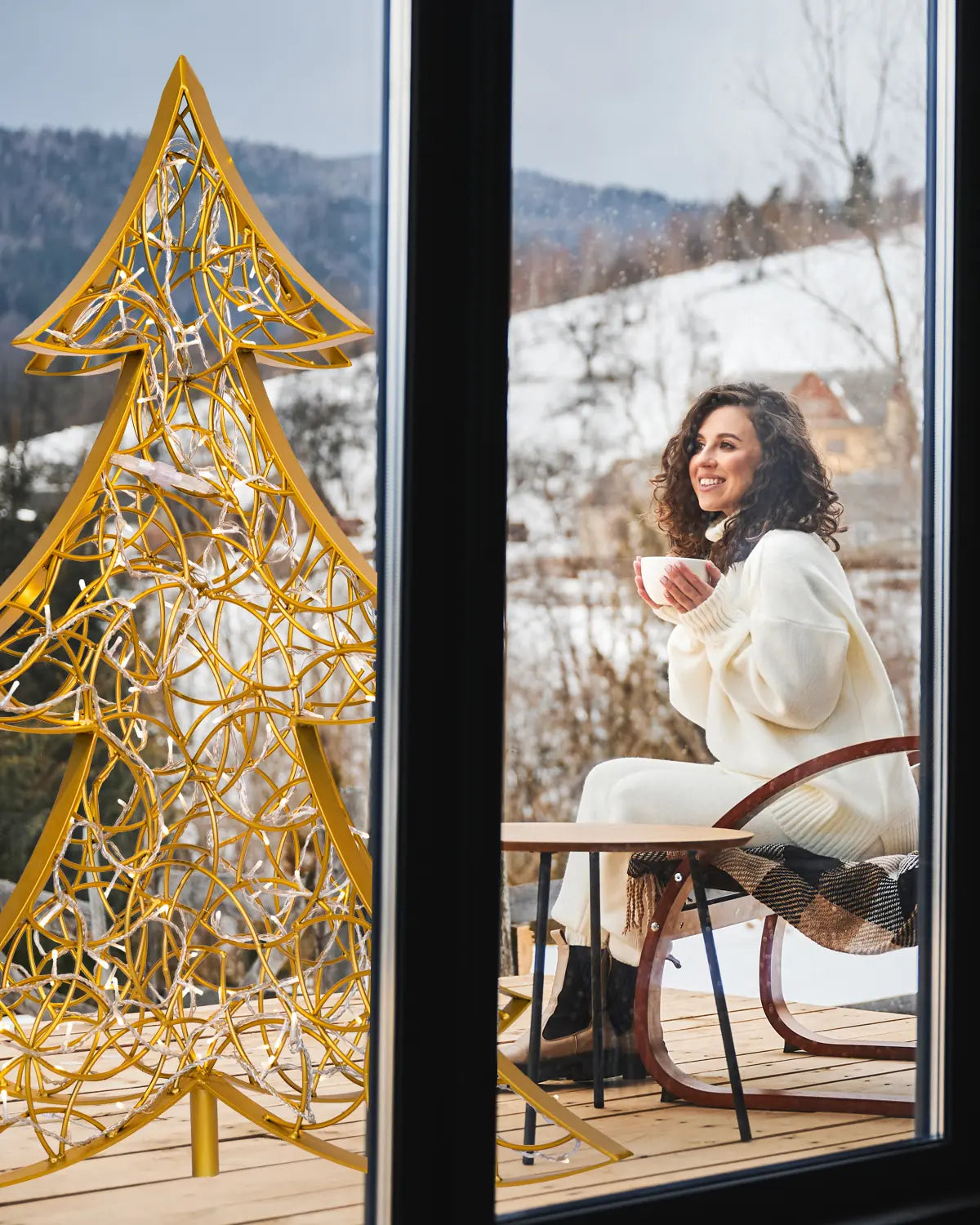 Goldener Alganic-Lichtbaum auf verschneiter Holzterrasse – Blick durch Fensterscheibe auf gemütliche Winterstimmung mit Bergpanorama.