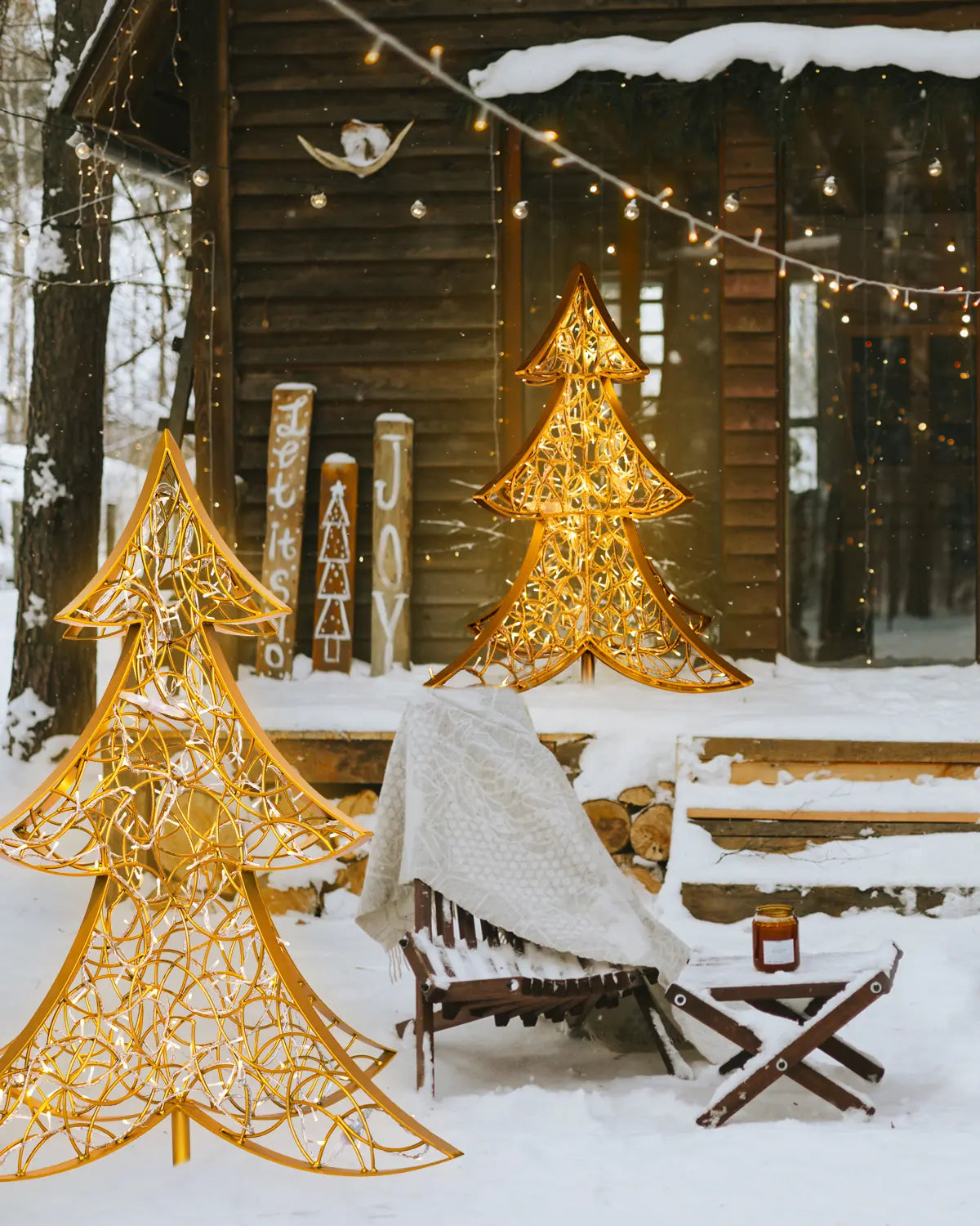 Zwei goldene Weihnachtsbäume von Alganic vor einer verschneiten Hütte – stimmungsvolle Lichtobjekte für winterliche Außenbereiche mit Holzarchitektur.