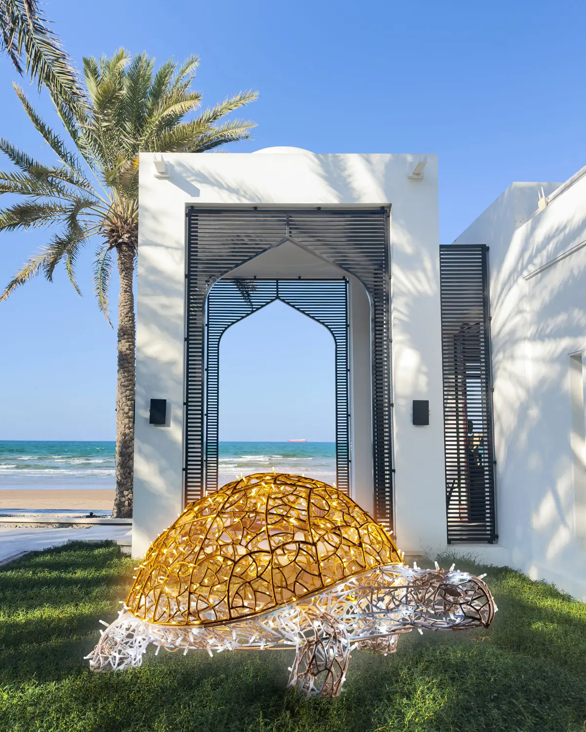 Leuchtende Schildkröten-Skulptur aus Aluminium im Gras vor modernem Beachresort – taghelle Installation mit Meeresblick.