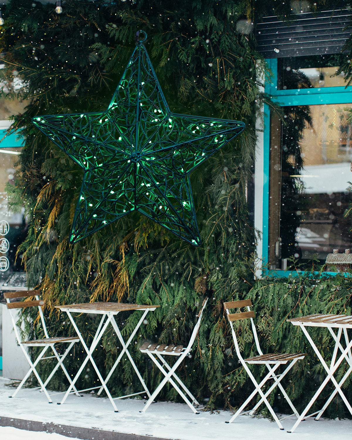 Grüner leuchtender Stern aus Draht vor einer Wintertanne – stimmungsvolle Dekoration im Außenbereich eines Straßencafés.